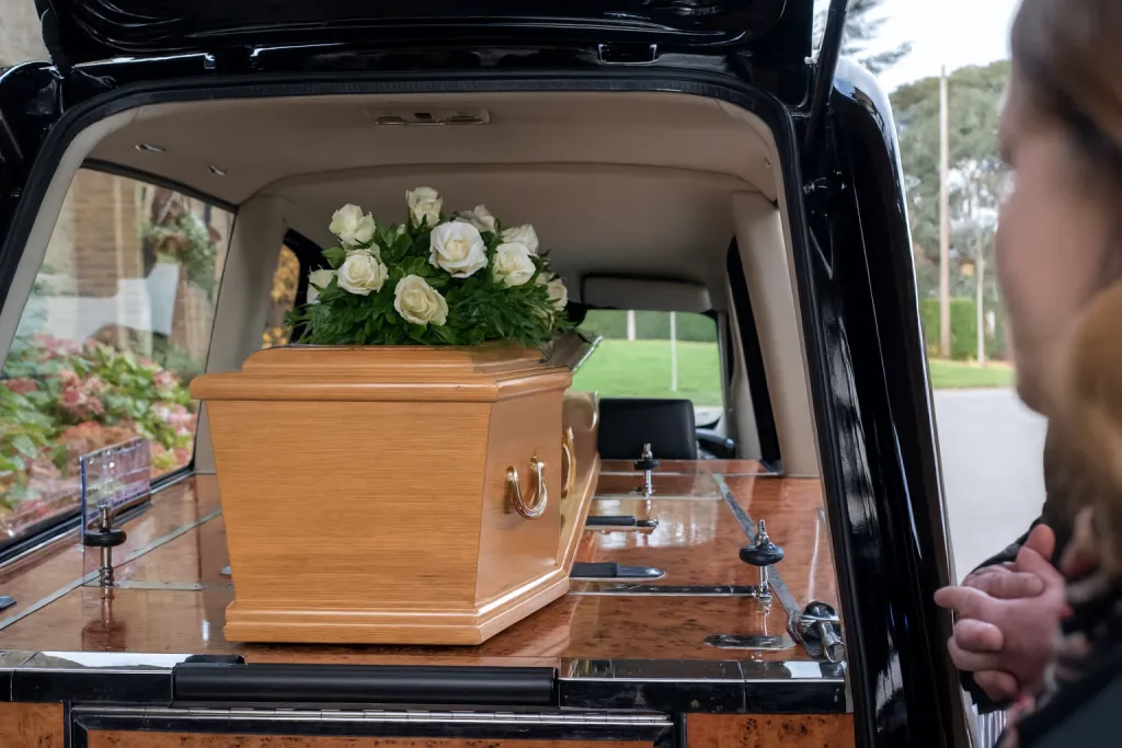 Cercueil en bois clair surmonté de roses blanches installé à l'intérieur d'un corbillard noir